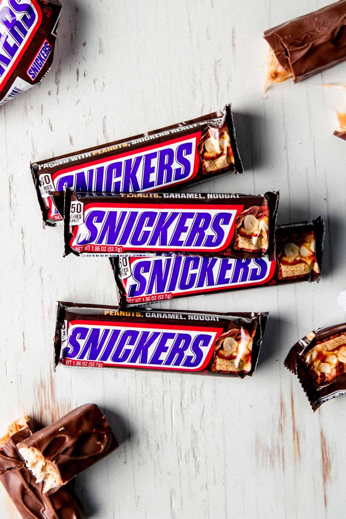 Snickers candy bars on white wooden surface, some unwrapped and broken apart.