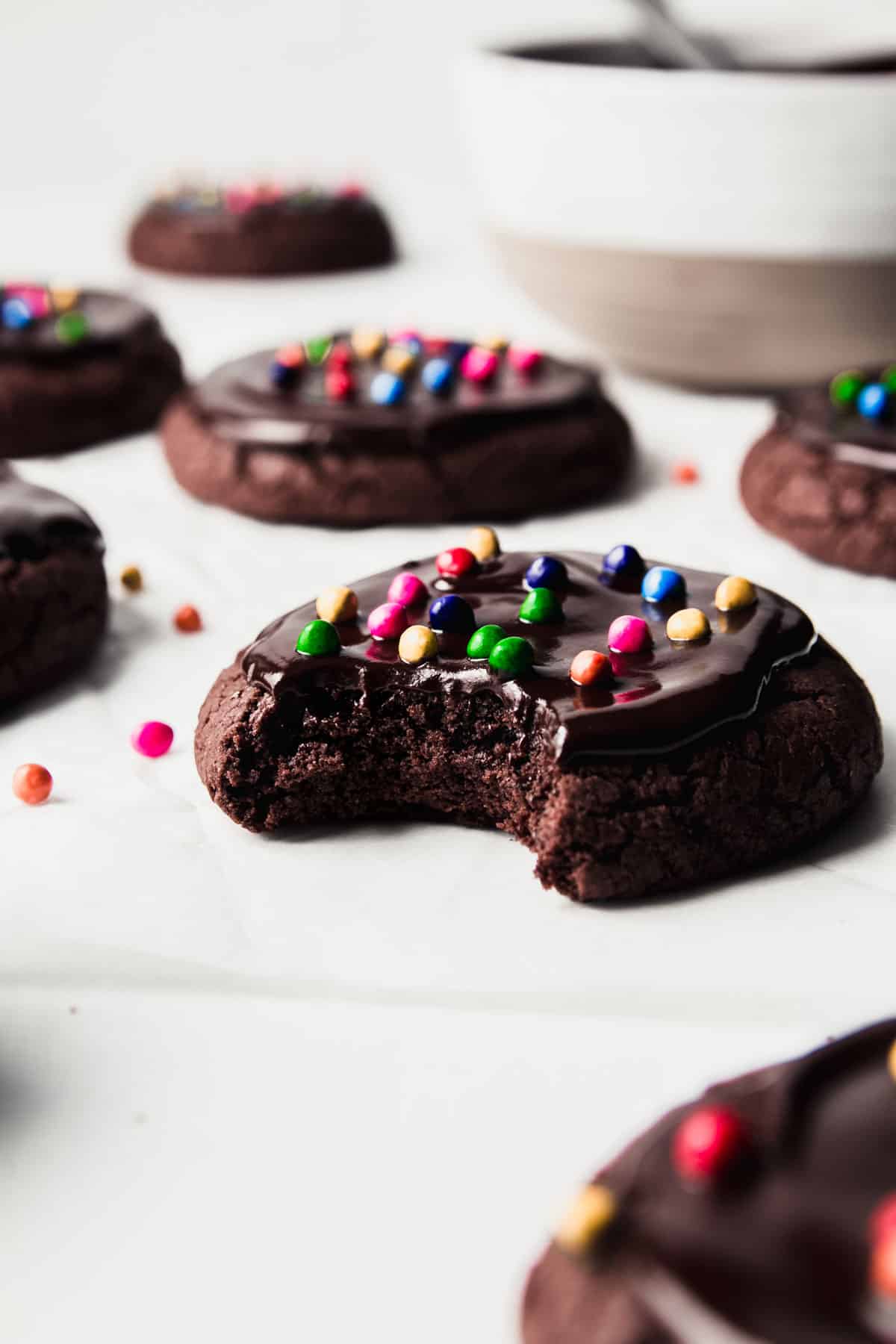 A rich brownie cookie topped with ganache and candy sprinkles, a bite taken showing interior texture.
