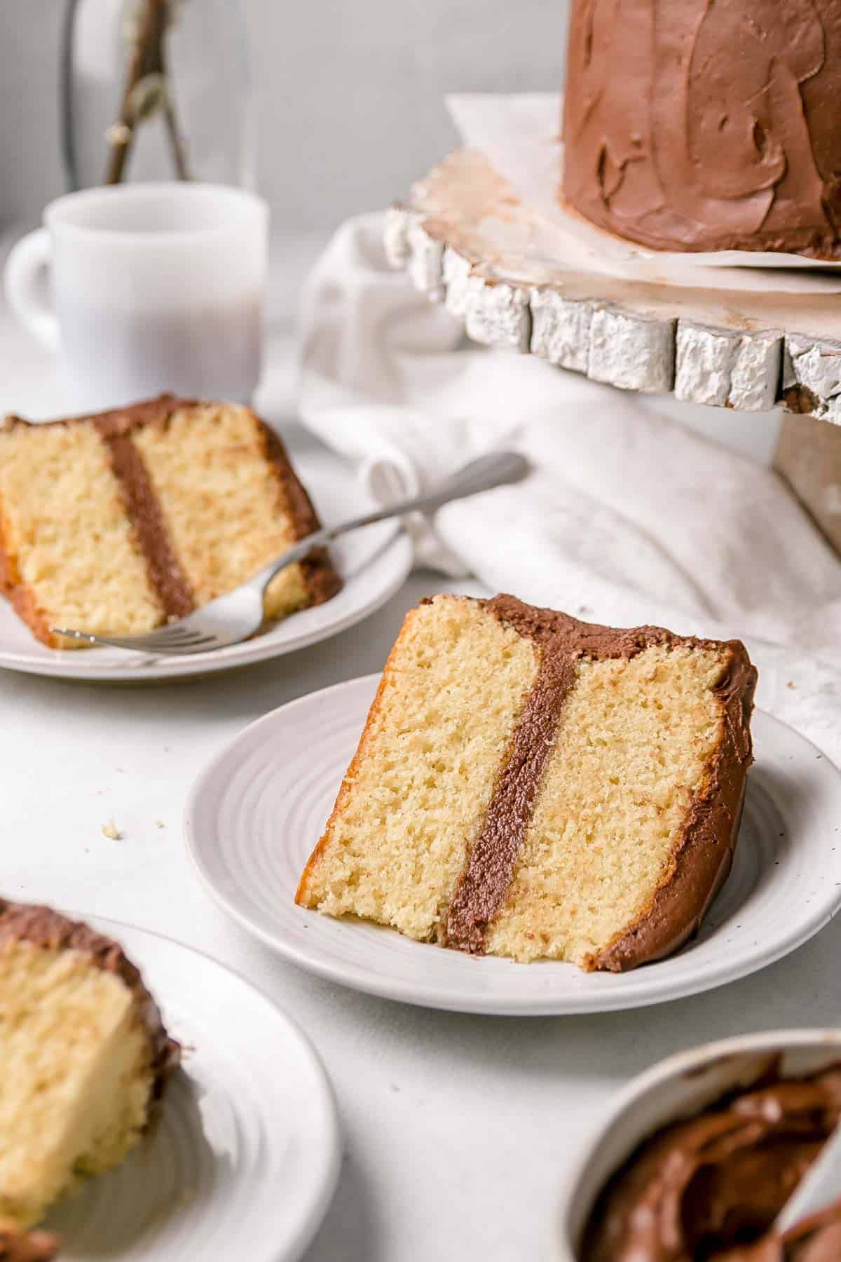 A slice of gluten free yellow cake with chocolate frosting on a small white plate.