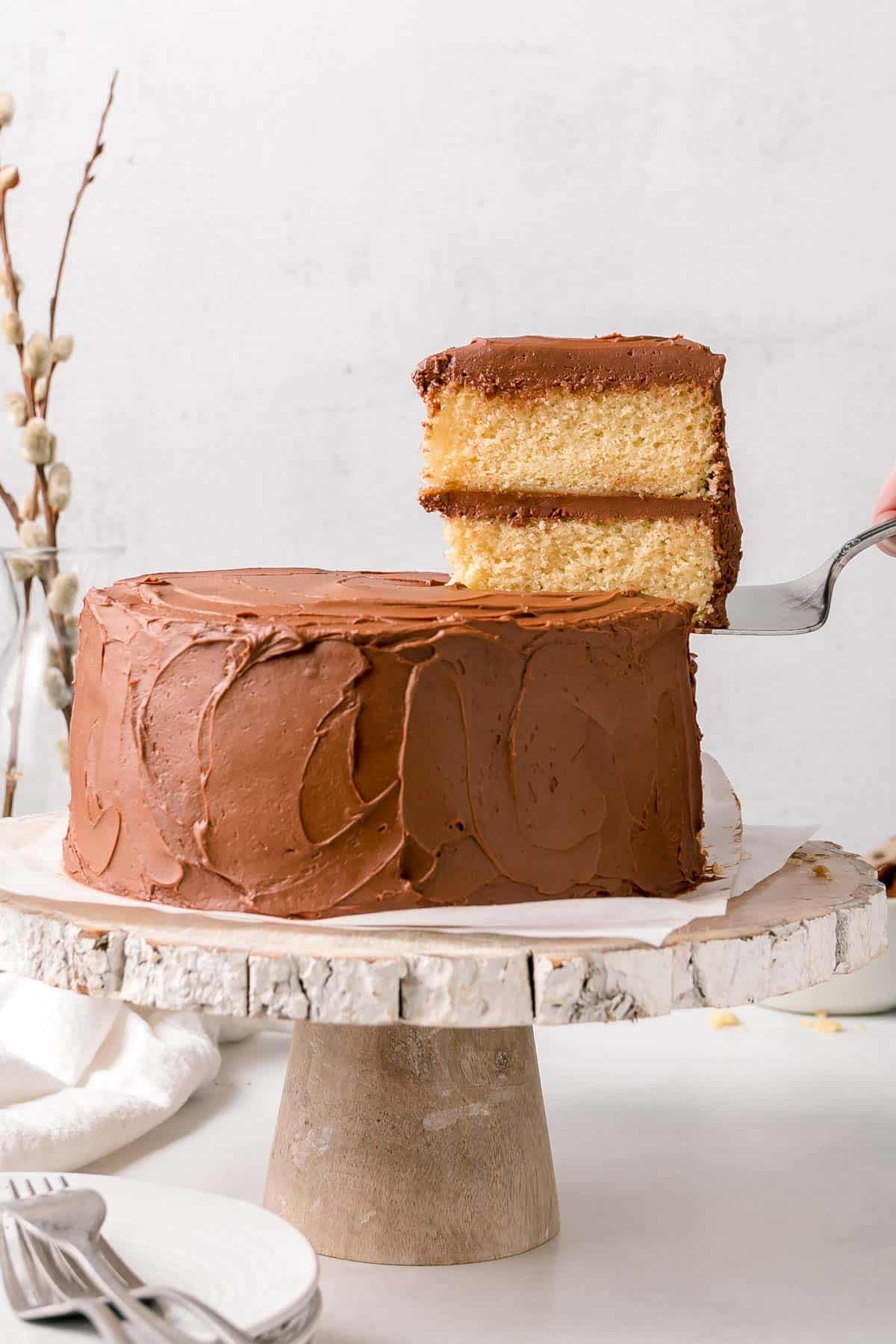 Two layer gluten free vanilla cake with chocolate frosting slice on serving spatula next to whole cake.