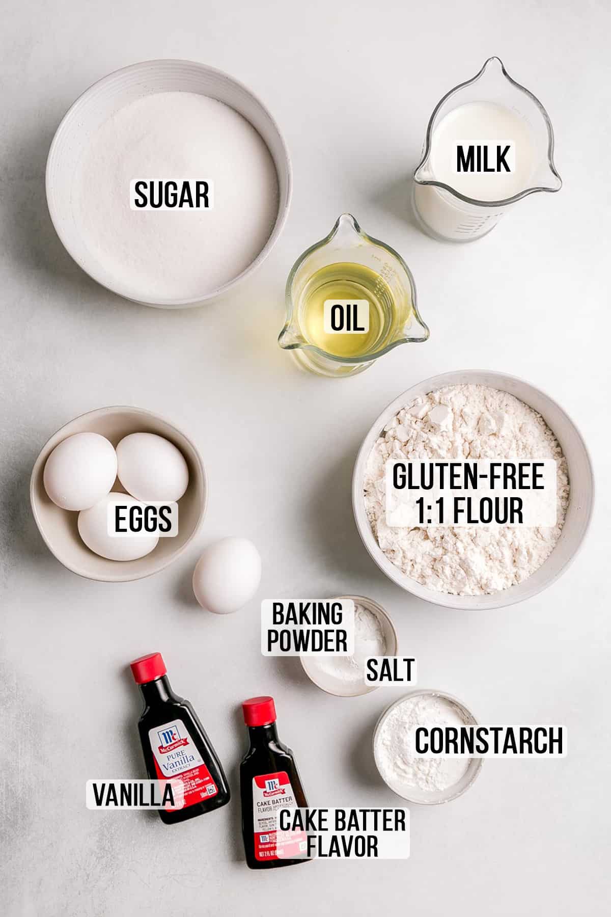 Ingredients for gluten-free yellow vanilla cake measured in bowls on white surface. 