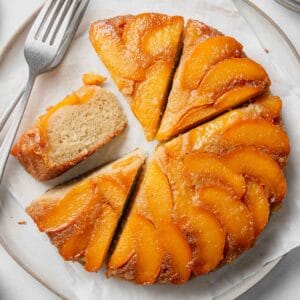 A round gluten-free peach upside down cake cake with peaches arranged on top with some slices cut, one on its side.