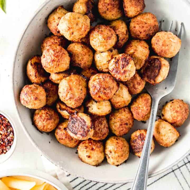 A bowl full of gluten-free baked chicken meatballs on white wooden surface next to a bowl of pasta.