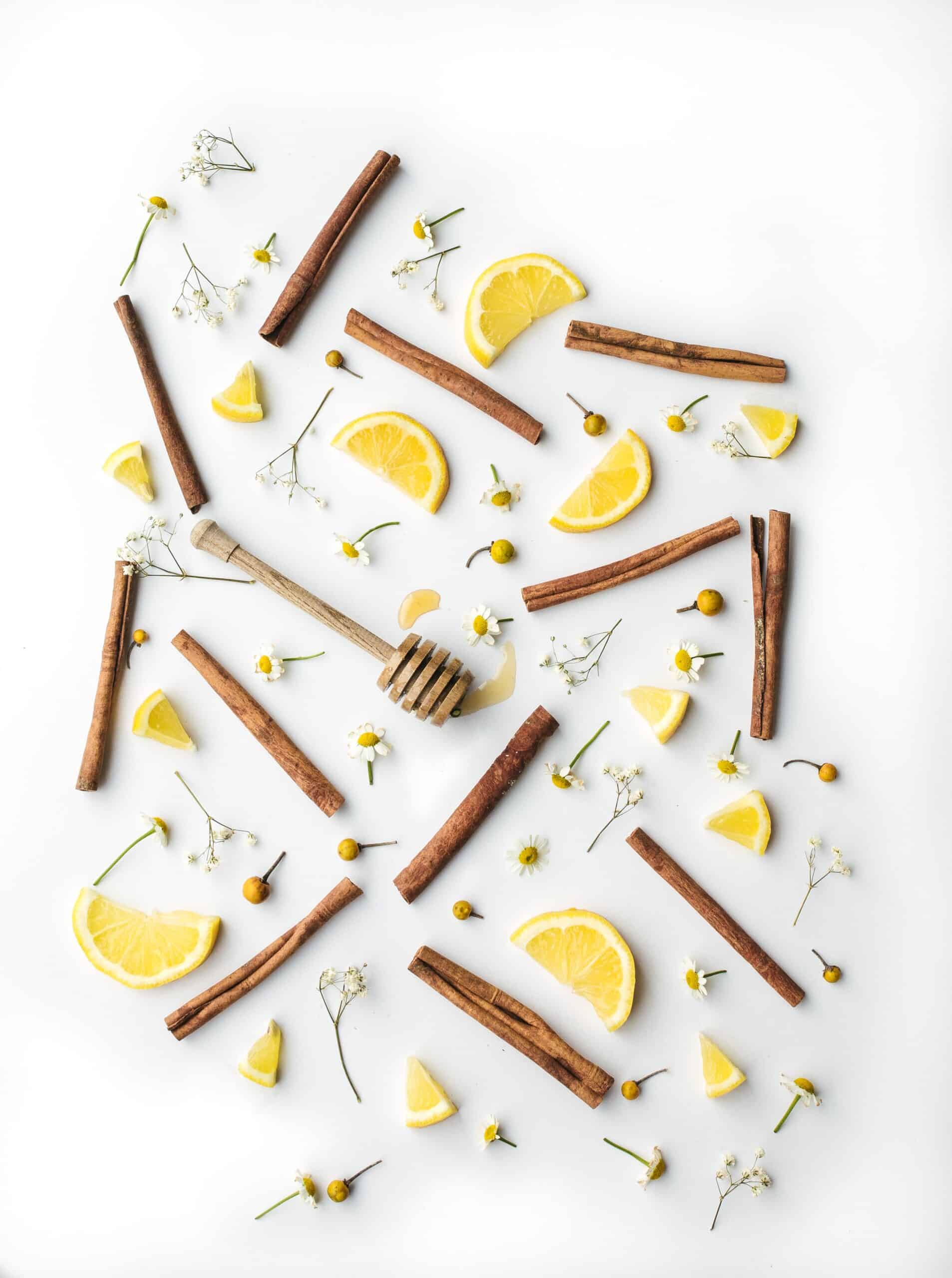 Honey, lemon wedges, cinnamon sticks, and small white flowers scattered on white surface.