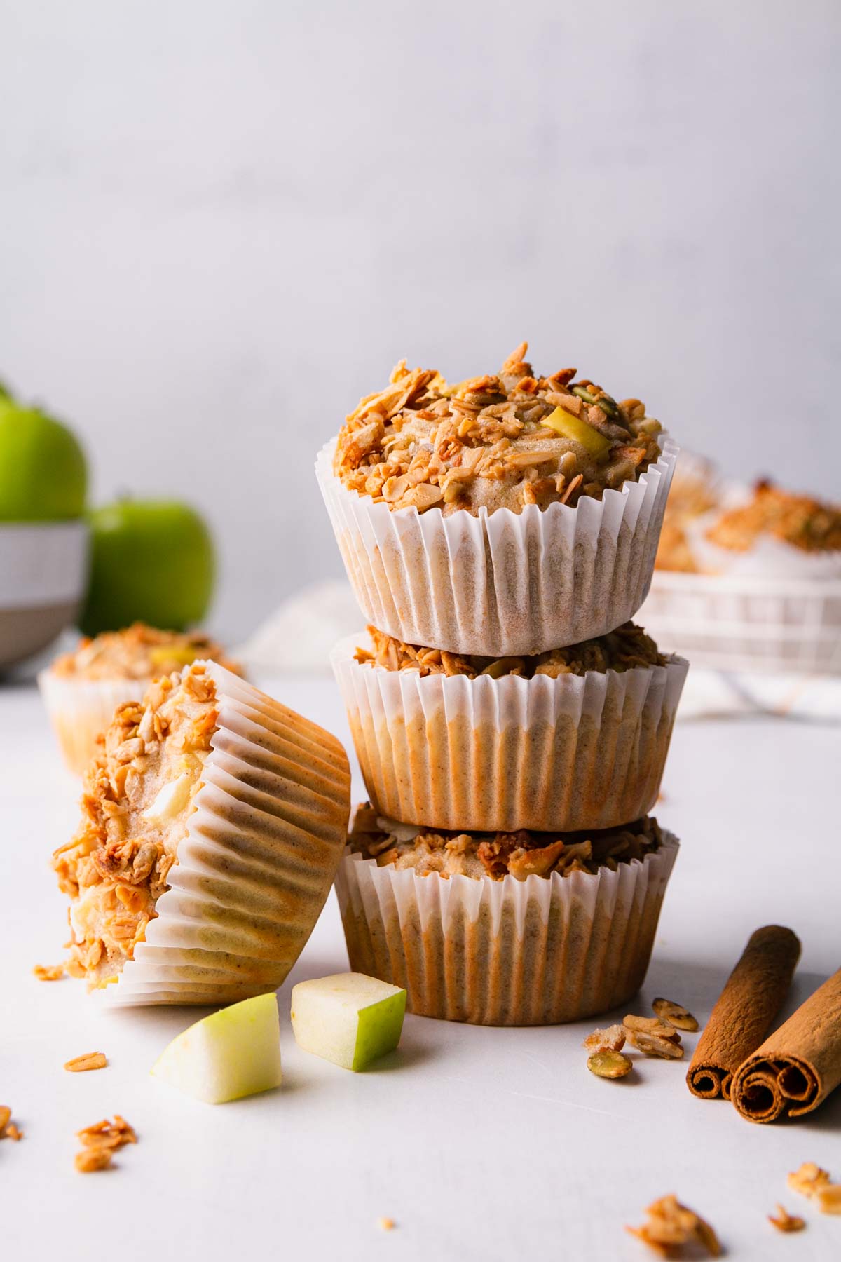 A stack of three gluten-free apple granola muffins with one more leaning on the side of the stack. Surrounded by diced apples and cinnamon sticks.