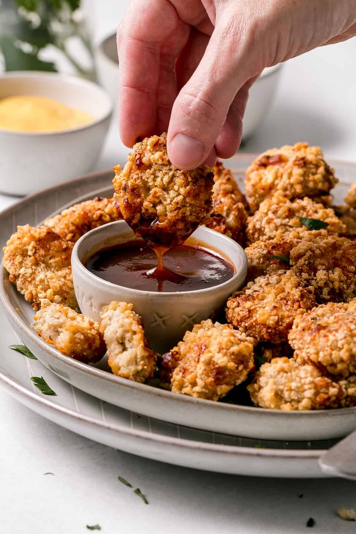 Chicken nugget dunked in barbecue sauce next to plate full of nuggets. 