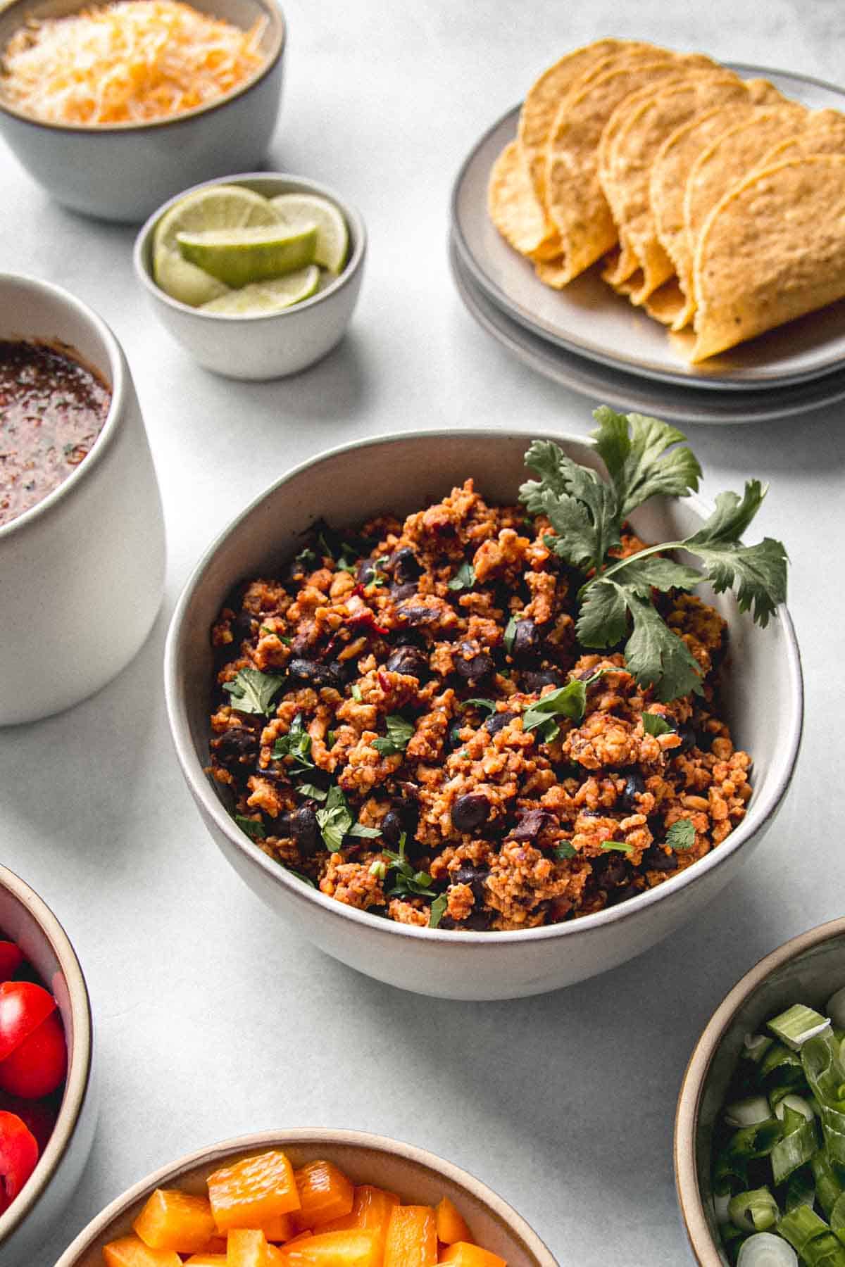 Ground chicken taco meat in a bowl topped with cilantro and surrounded by bowls of taco fixings.