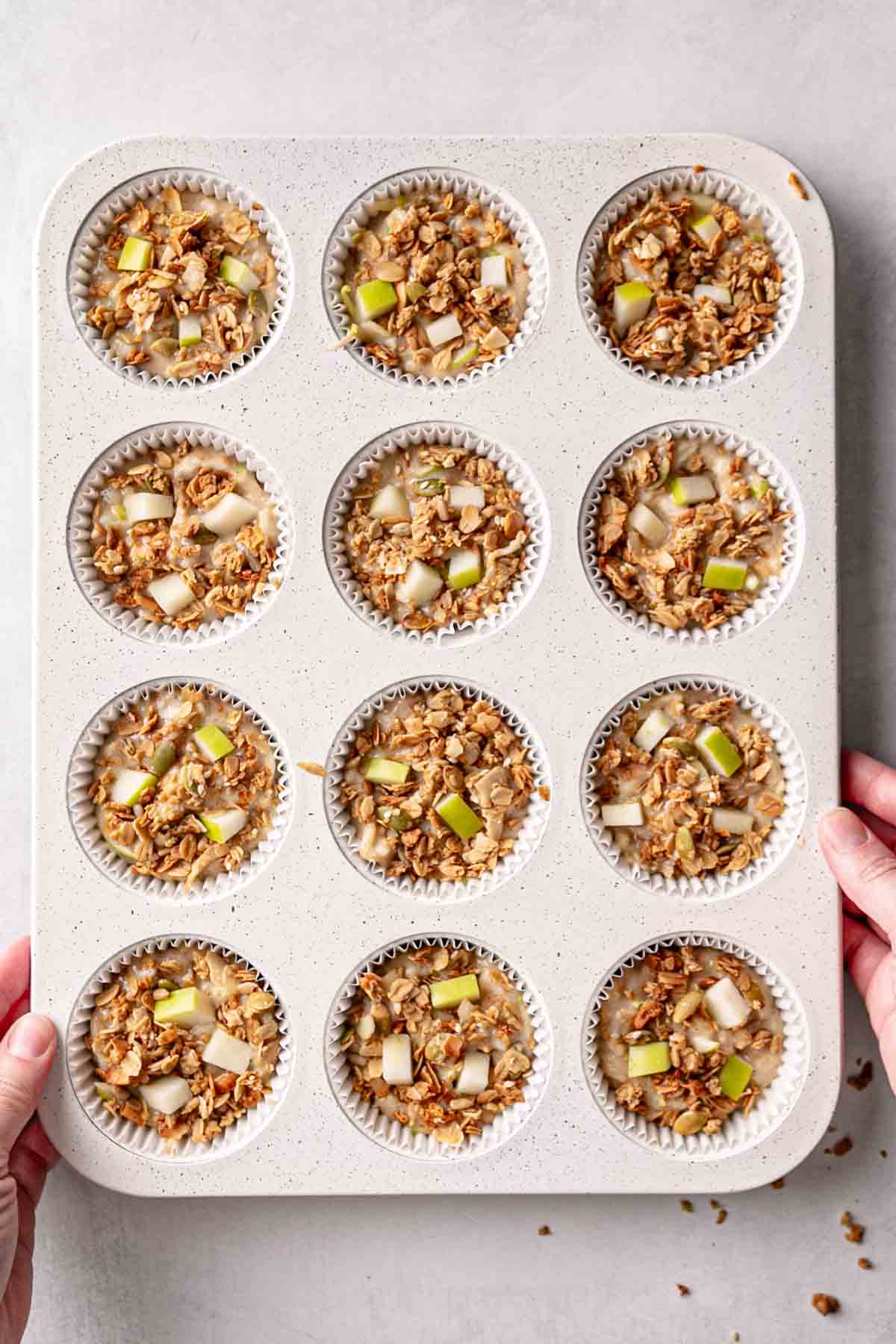 Muffin batter in white muffin tin, granola and diced apples pressed on top of batter.