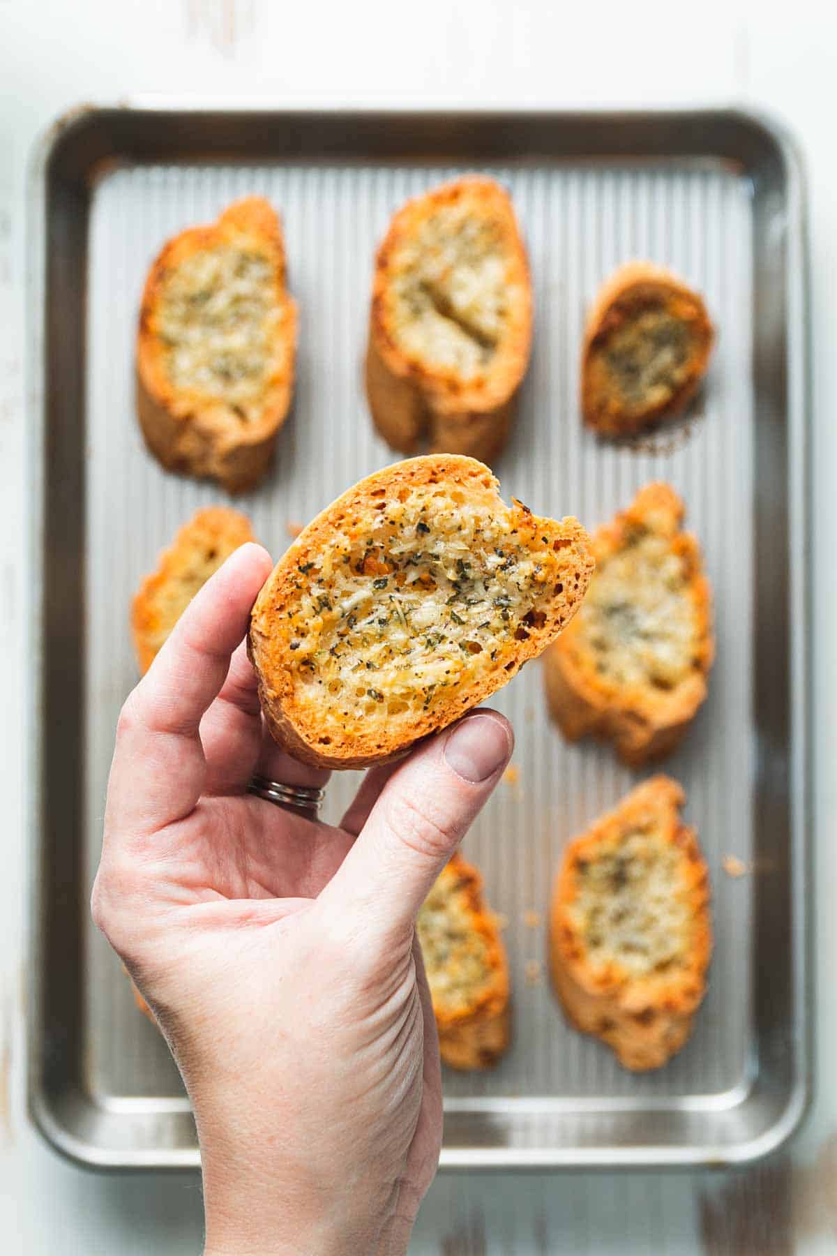 A hand holding a piece of golden brown garlic bread over a baking tray with the rest of the garlic bread slices.