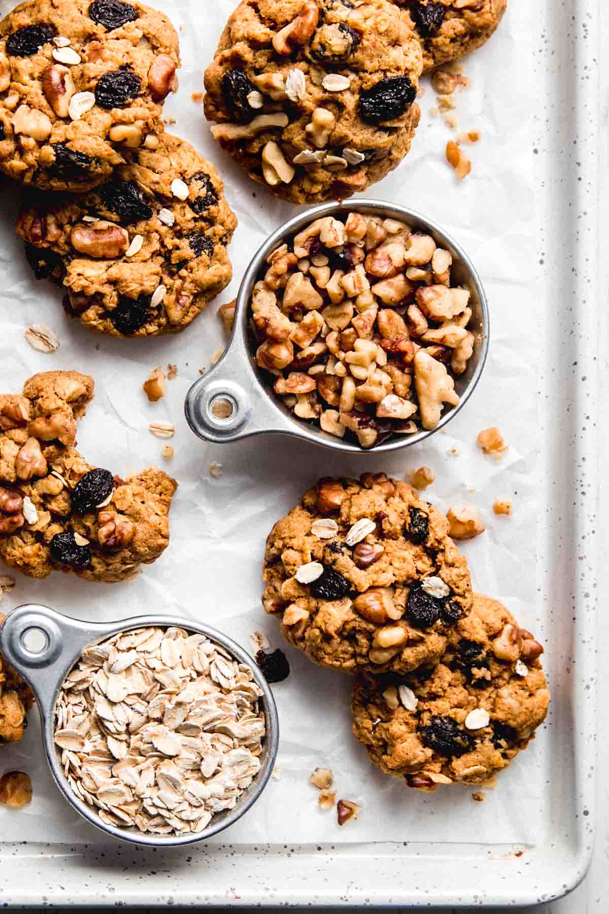 A speckled baking sheet filled with cookies and measuring cups containing oats or walnuts.