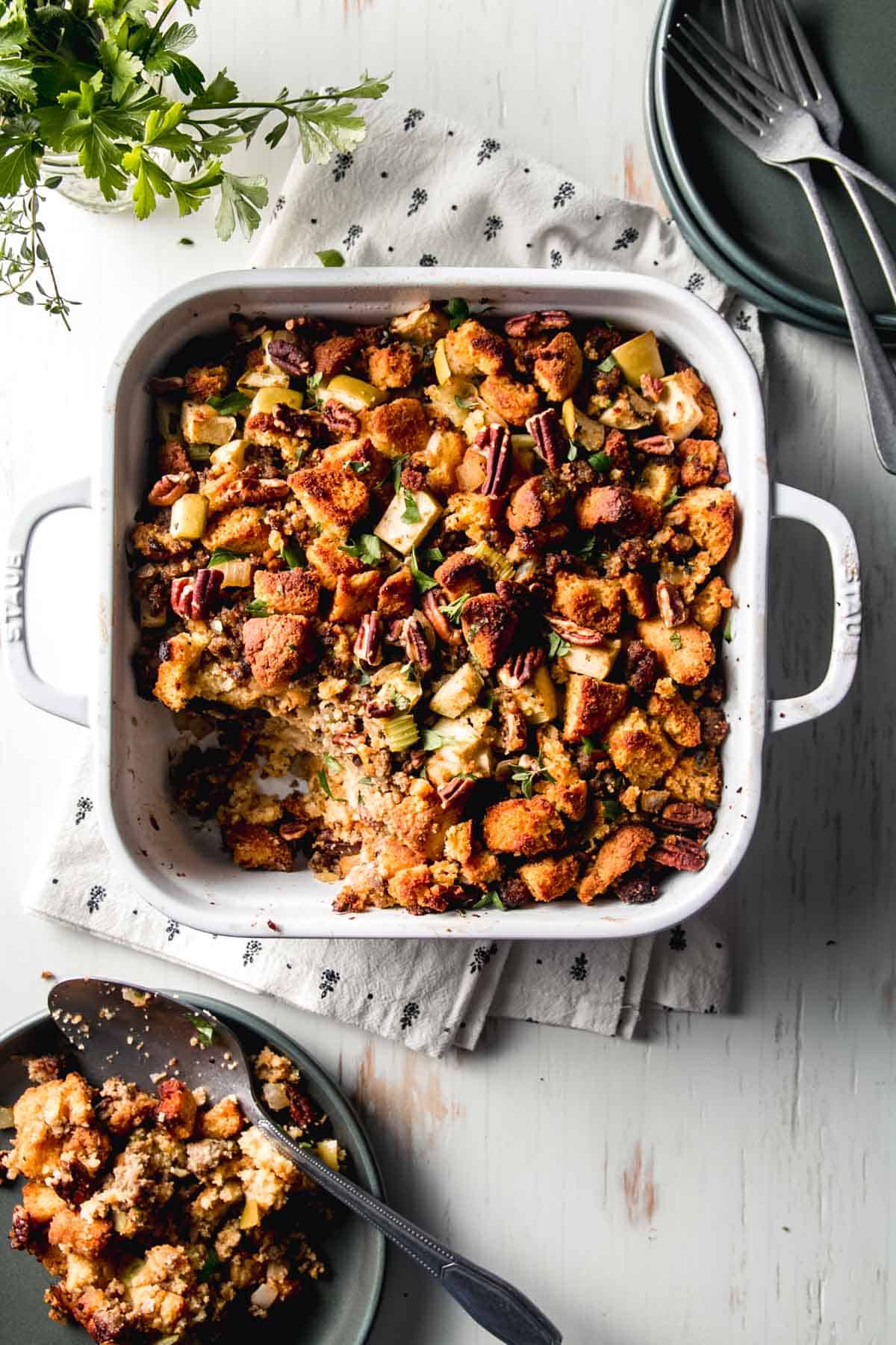 Gluten-free cornbread stuffing in a white casserole dish next to green plates, fresh herbs, and forks.