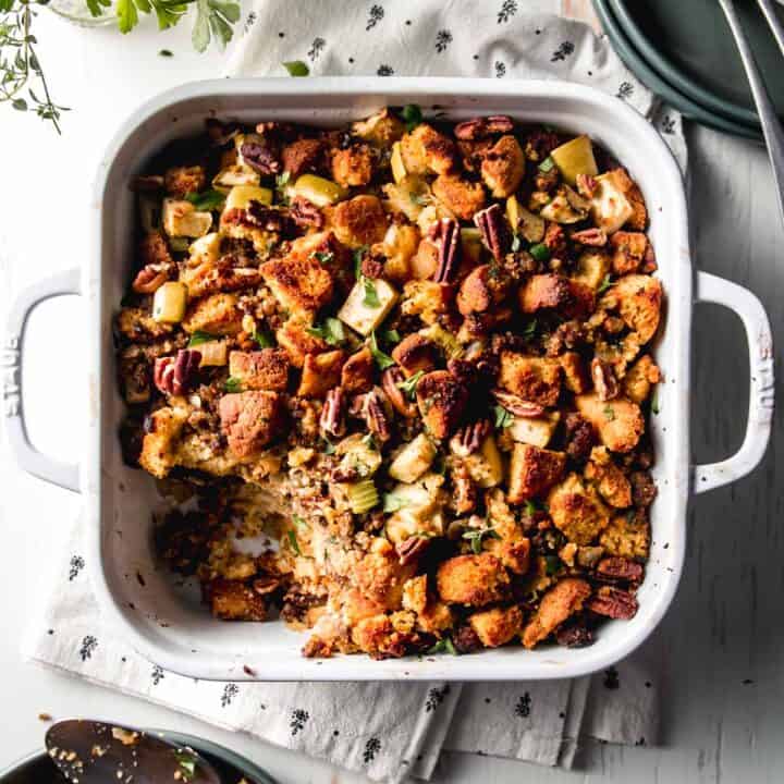 Gluten-free cornbread stuffing in a white casserole dish next to green plates, fresh herbs, and forks.