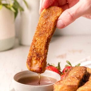 A french toast stick with syrup dripping down over a small bowl.