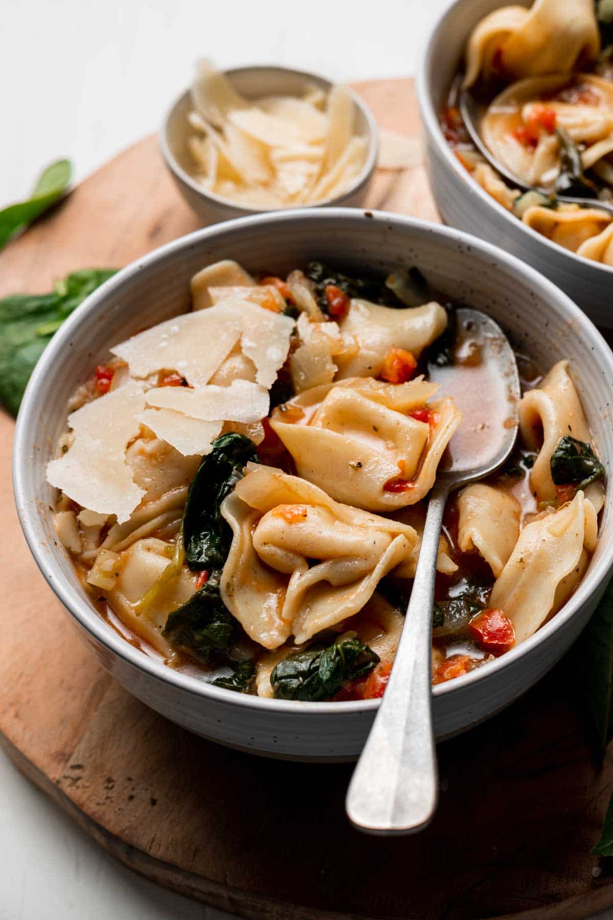 Tortellini soup in bowl with spoon, spinach and parmesan cheese on top.