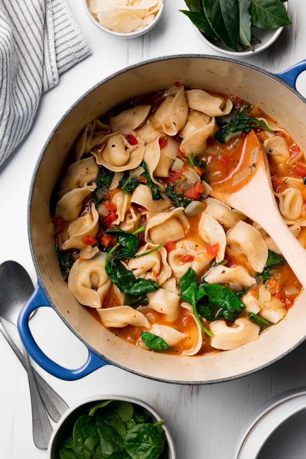 Gluten free spinach tortellini soup in large blue pot next to baby spinach and shaved parmesan cheese.