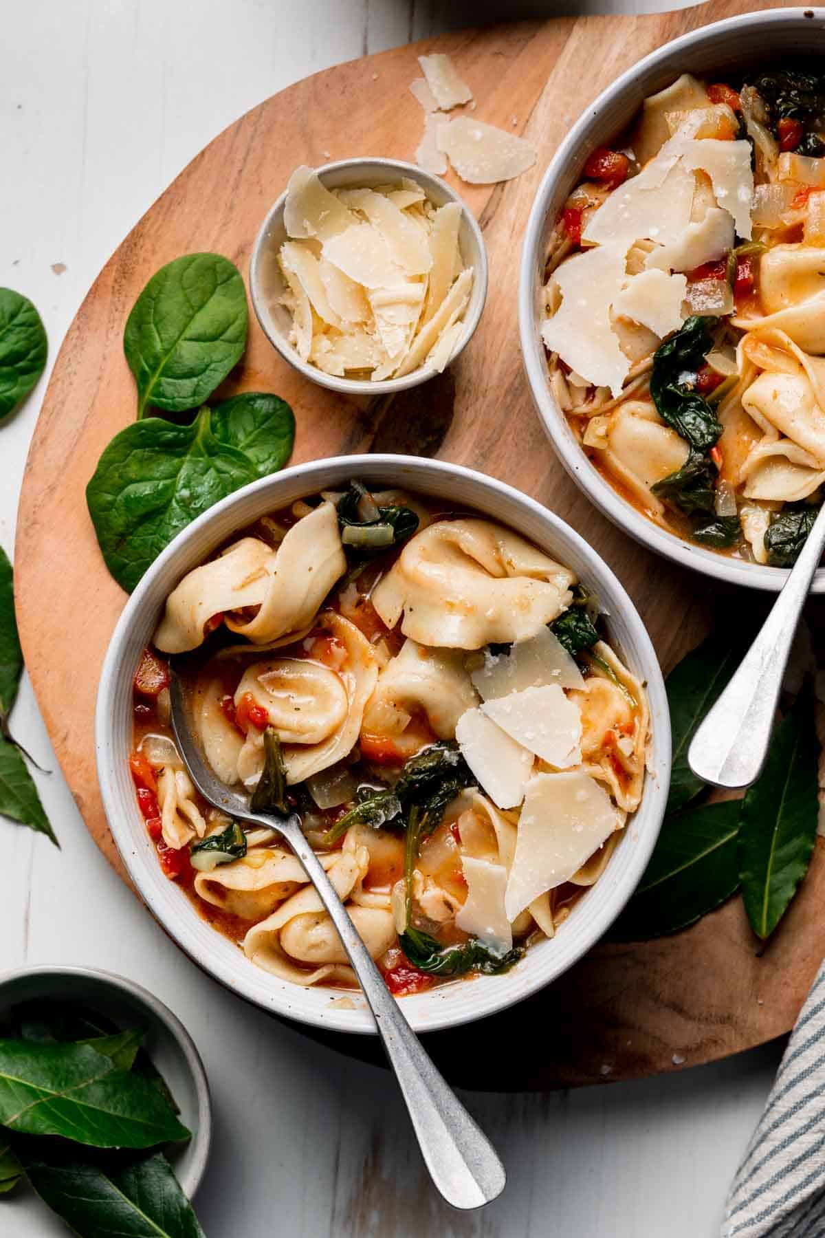 Two bowls of gluten free tortellini soup next to shaved parmesan and baby spinach on a wooden surface.