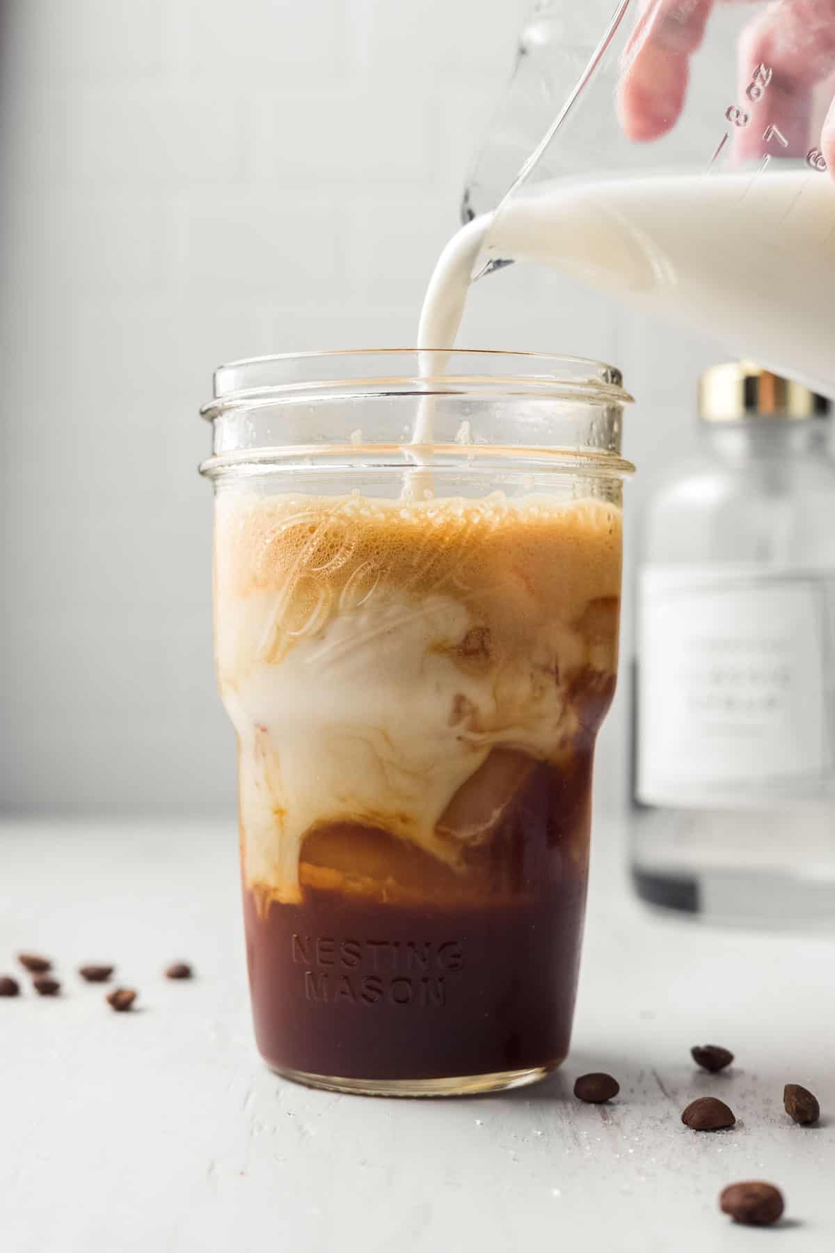 Pouring milk into mason jar that has ice and shaken espresso in it, bubbles on top.