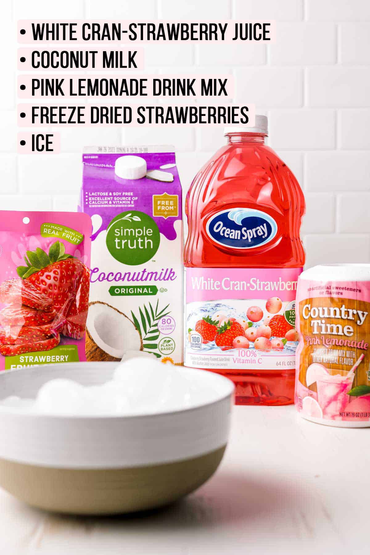 A bottle of ocean spray white-cran-strawberry juice, next to a carton of coconut milk, a package of freeze dried strawberries, country time lemonade mix, and a bowl of ice.