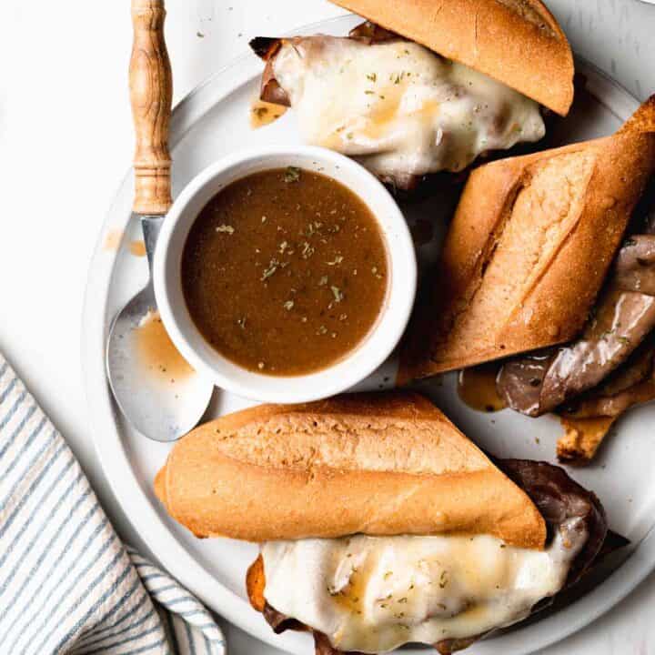 Gluten free French dip sandwiches with melted cheese and au jus dips on top next to a cup of au jus.