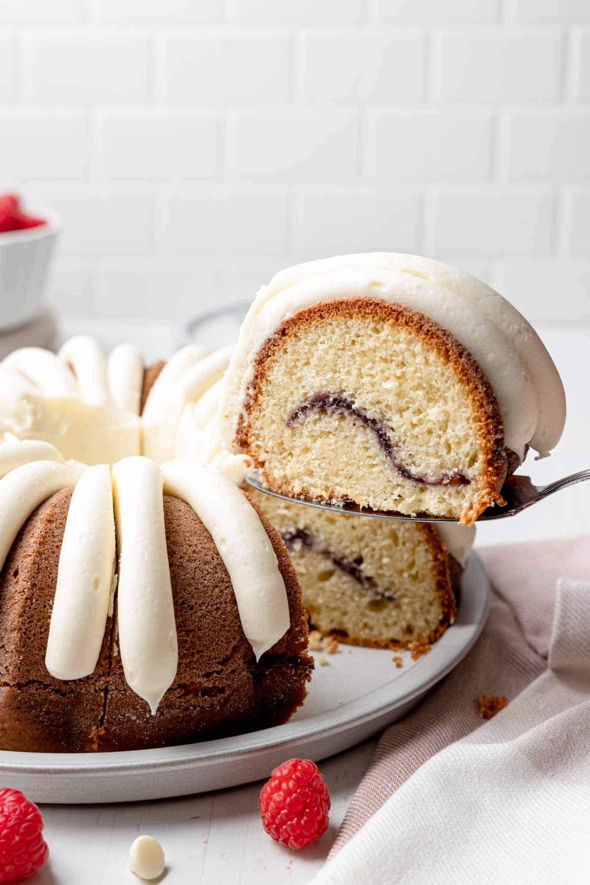 A slice of bundt cake on a serving spatula is lifted from the rest of the cake showing interior of the cake and swirl of raspberry. 