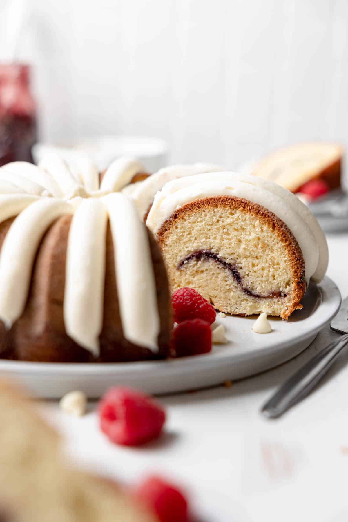 Gluten free white chocolate bundt cake slice with a raspberry swirl in the center and lines of cream cheese frosting piped on top.