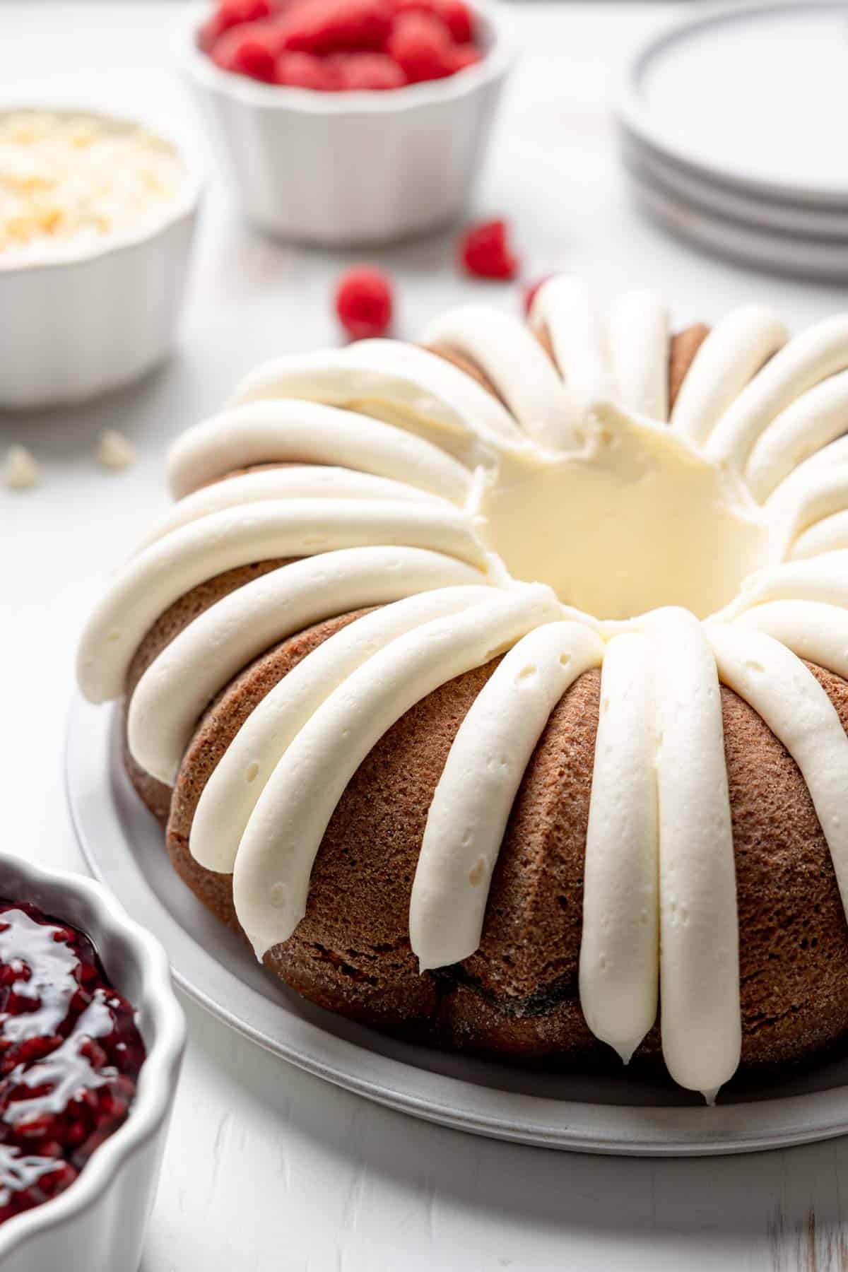 Bundt cake with cream cheese icing piped around the cake next to raspberries and white chocolate chips.