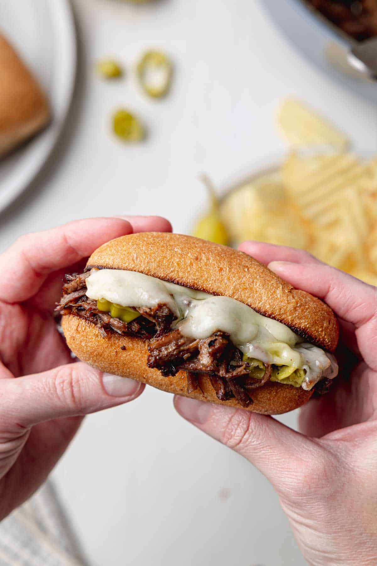 Two hands holding a shredded Italian beef sandwich on a gluten-free ciabatta roll with gooey melted cheese.