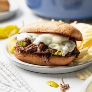 Italian beef sandwich with melted provolone cheese on small plate next to peperoncini peppers.