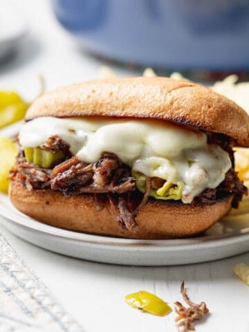 Italian beef sandwich with melted provolone cheese on small plate next to peperoncini peppers.