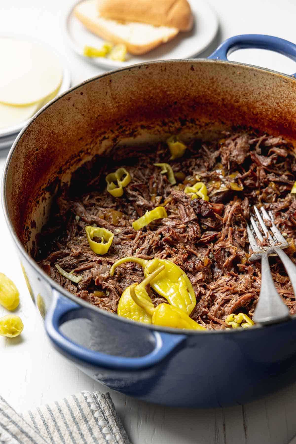 Italian beef shredded in a large blue pot with pepperoncini peppers on top.