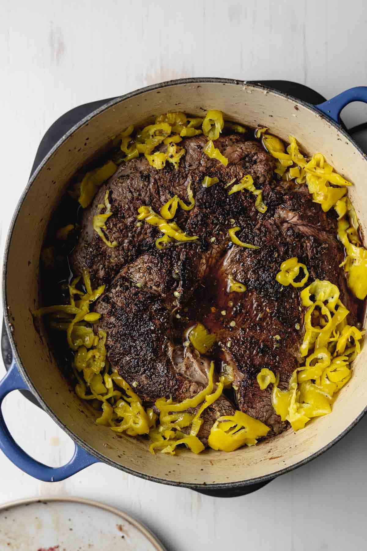 The beef in a large blue pot surrounded by pepperonccini peppers.