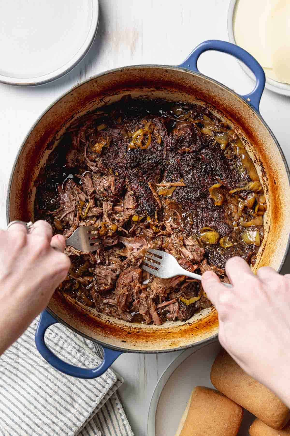 Two hands with forks shredding the beef in large blue pot.