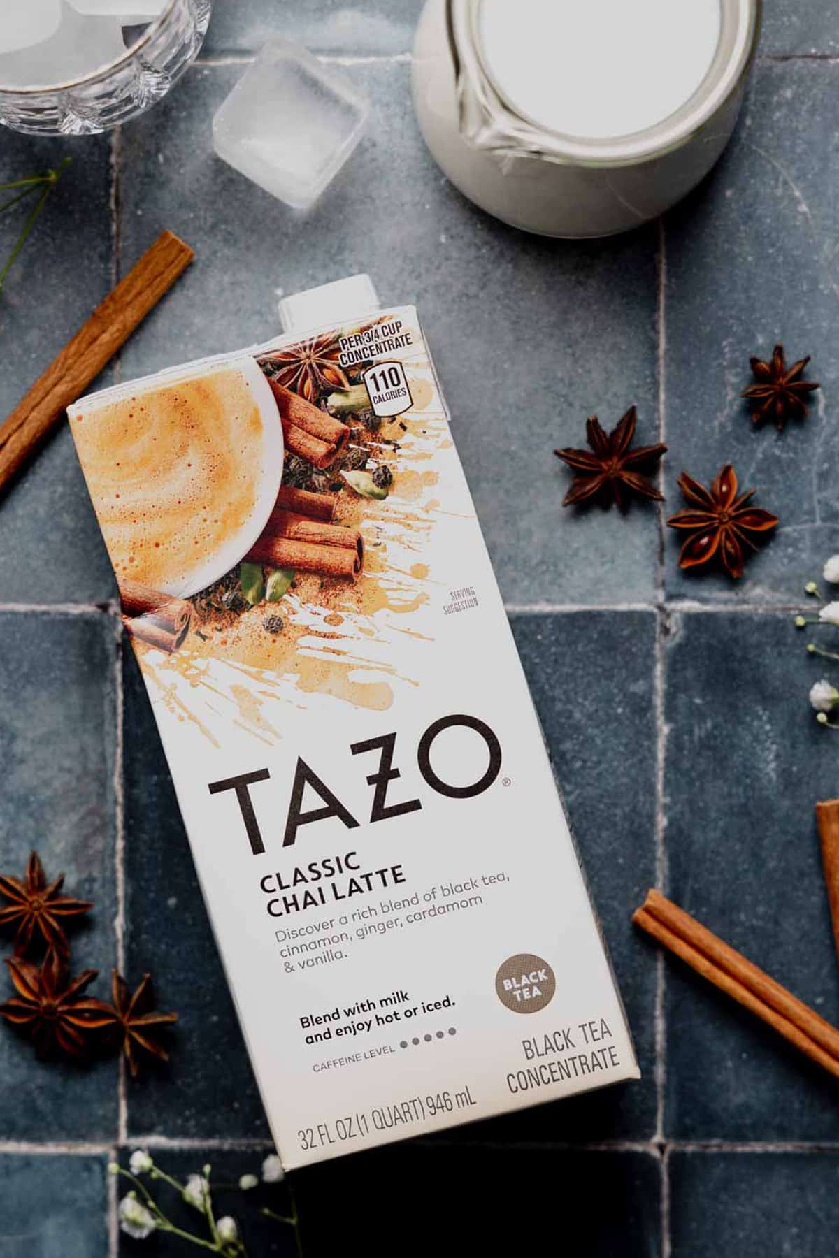 Tazo chai tea latte concentrate on blue tile surface net to a small pitcher of milk, star anise, and cinnamon sticks.