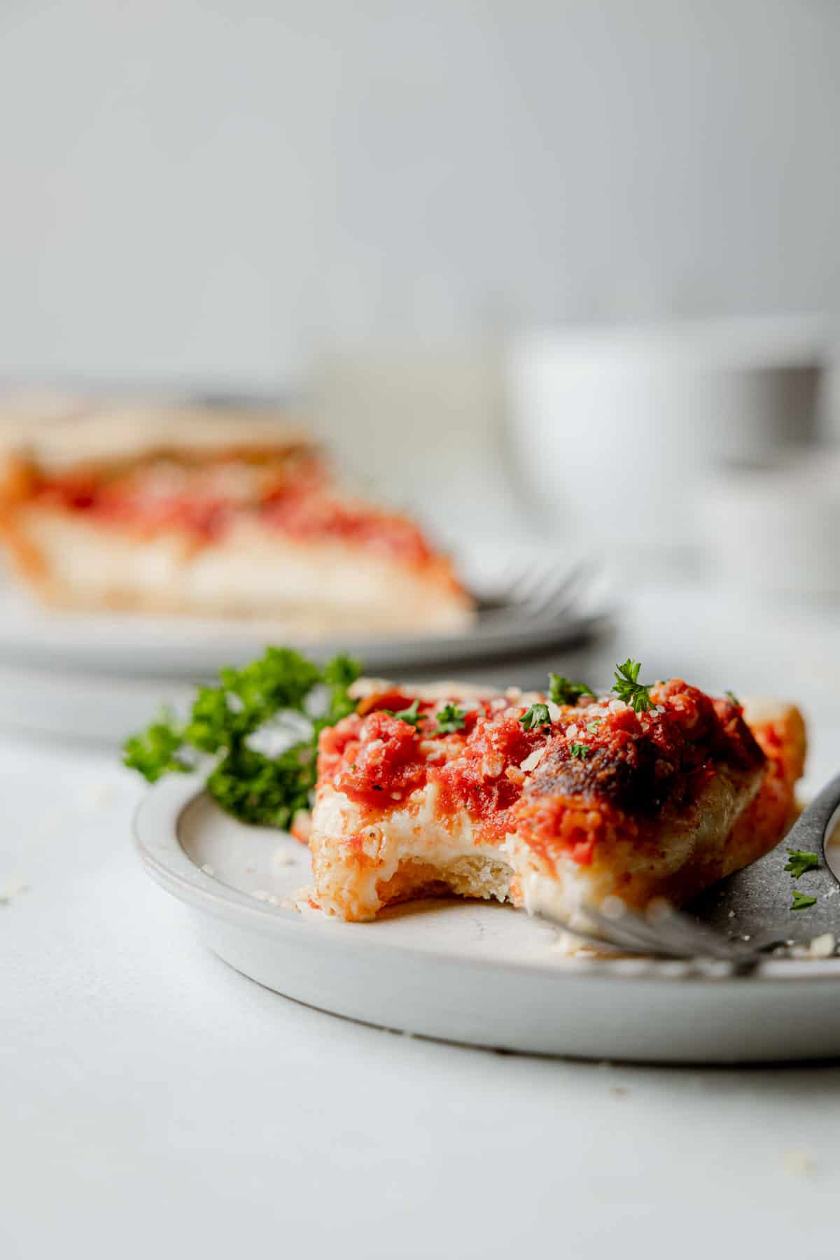 A slice of gluten free deep dish pizza on a small plate with bite taken, garnished with parsley.