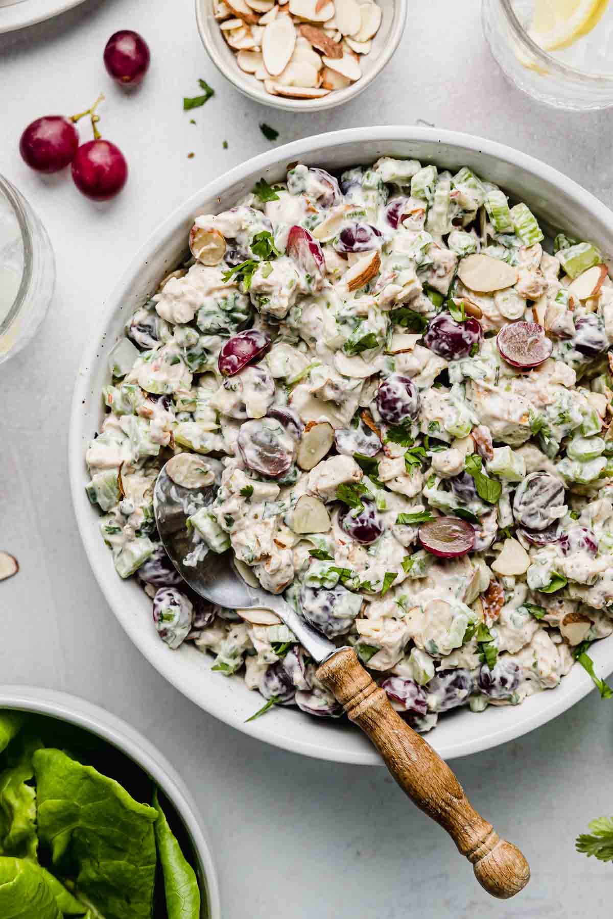 Chicken salad in large white bowl next to grapes, sliced almonds, and lettuce.