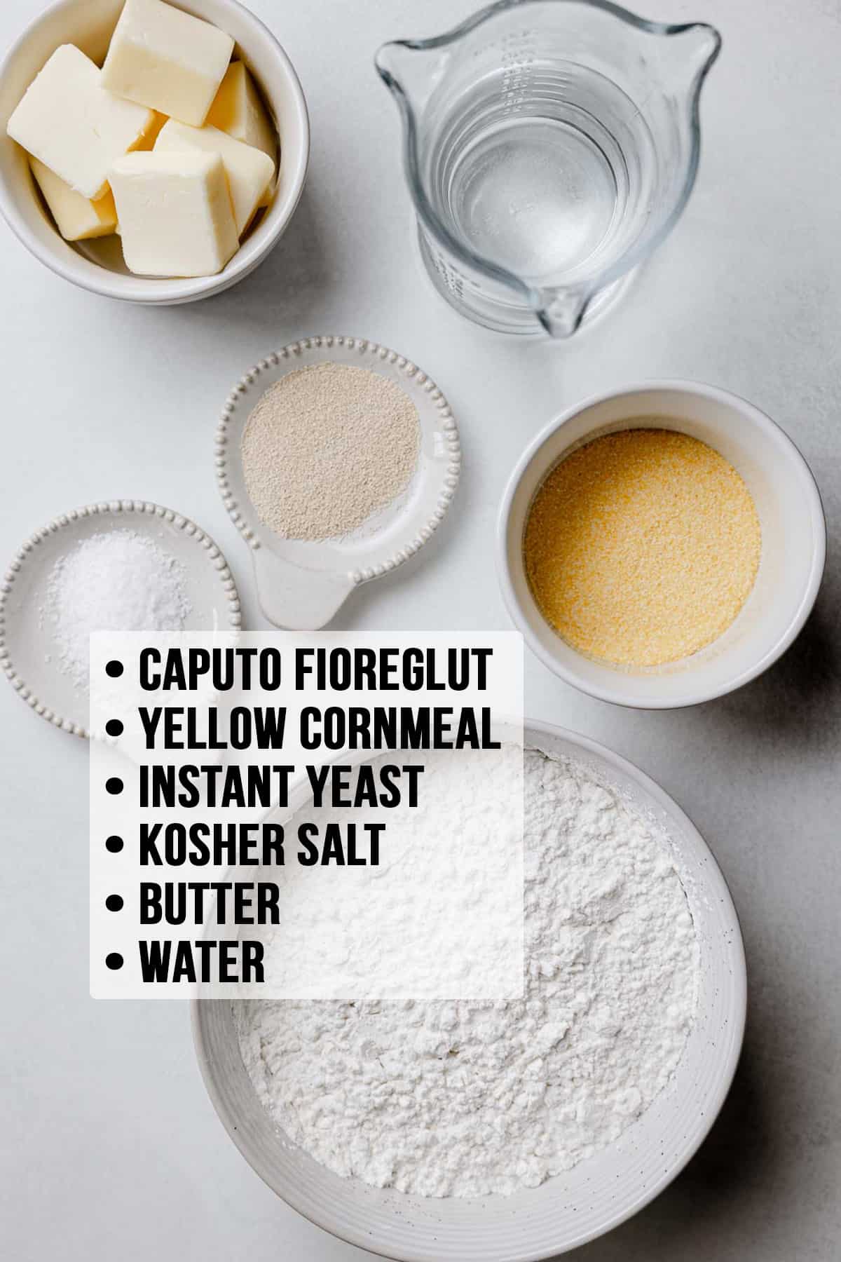 Captuo fioreglut, cornmeal, yeast, butter, salt, and water measured out into bowls on white surface.