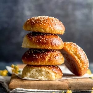 A stack of four gluten free burger buns with one leaning to the side, next to small yellow flowers on a wooden board.