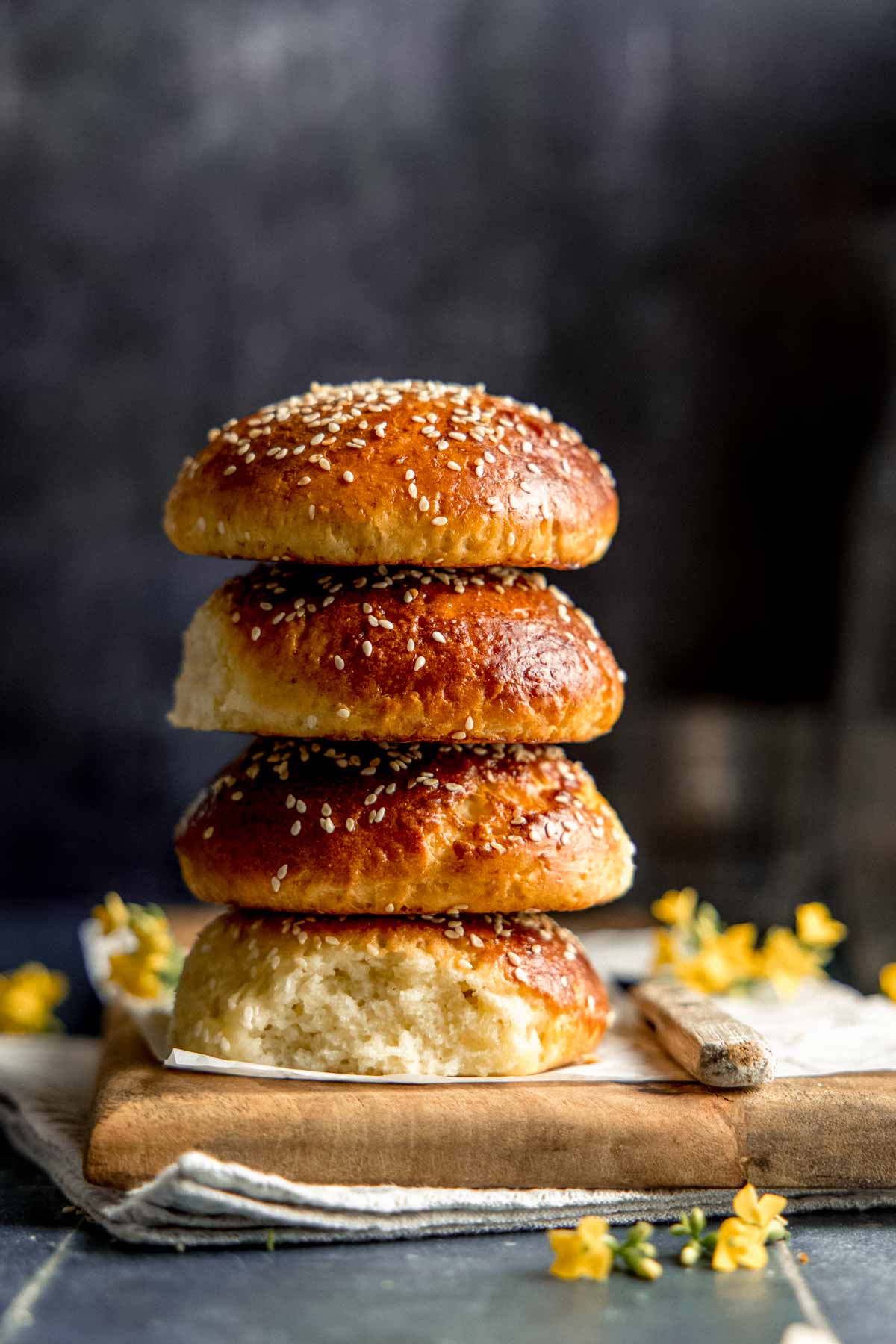 A stack of four gluten free burger buns with sesame seeds on top, next to small yellow flowers on a wooden board.