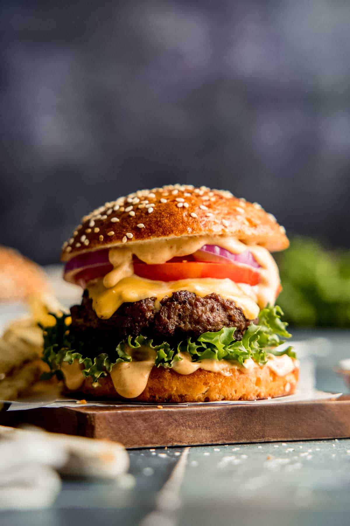 A gluten-free burger with lettuce, tomato, onion, and sauce on a gluten-free bun with sesame seeds.