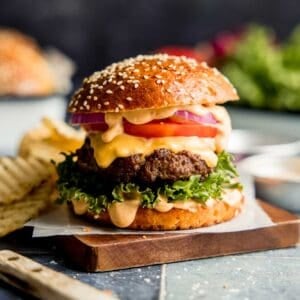 Hamburger topped with sauce, tomato, onion, and lettuce on wood cutting board.