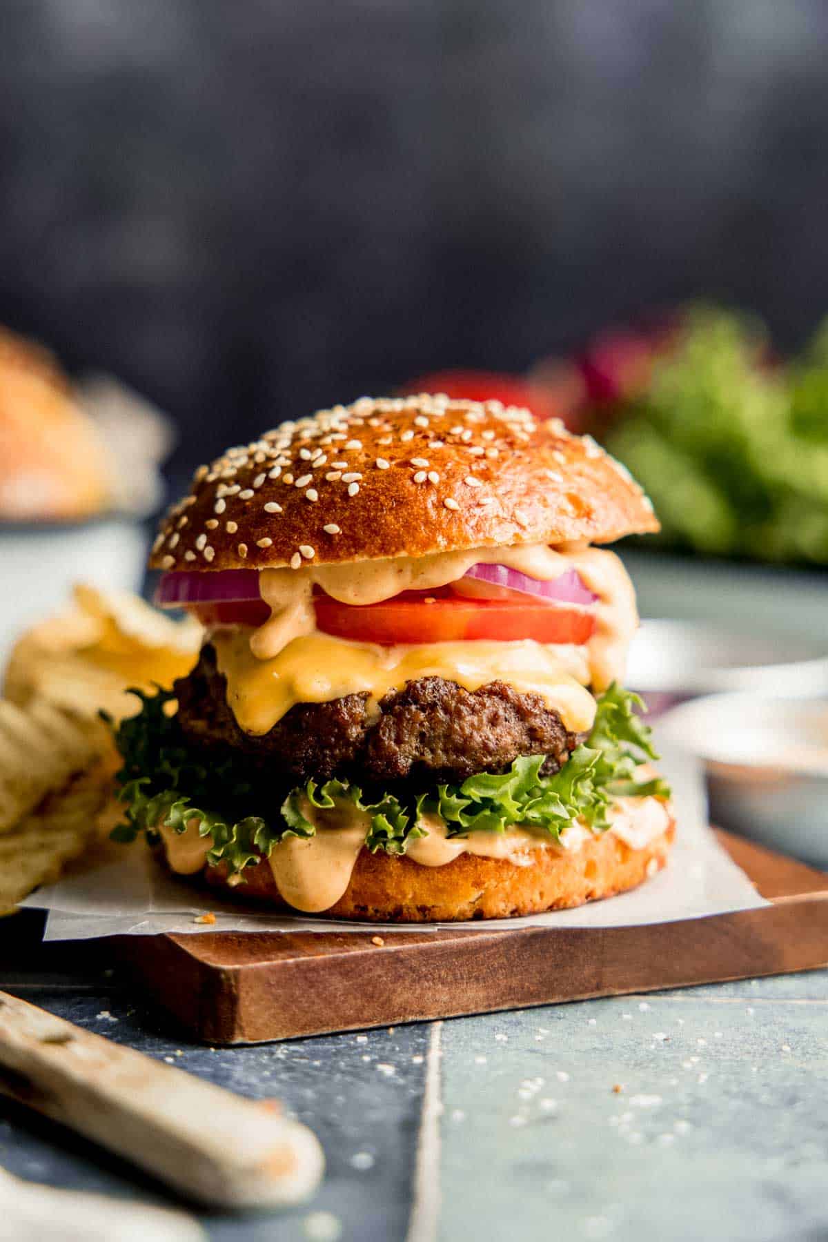 Burger with lettuce, tomato, onion, and sauce on a sesame seed bun, resting on wooden board.