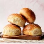 Side view of a stack of fluffy gluten-free hamburger buns with shiny, golden crusts and sesame seed topping, placed on a wooden board with a soft pink background.