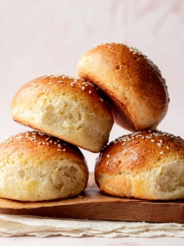 Side view of a stack of fluffy gluten-free hamburger buns with shiny, golden crusts and sesame seed topping, placed on a wooden board with a soft pink background.