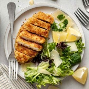 Gluten-free chicken cutlets sliced on plate next to small salad, shaved parmesan, and lemon wedges.