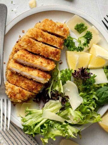 Gluten-free chicken cutlets sliced on plate next to small salad, shaved parmesan, and lemon wedges.