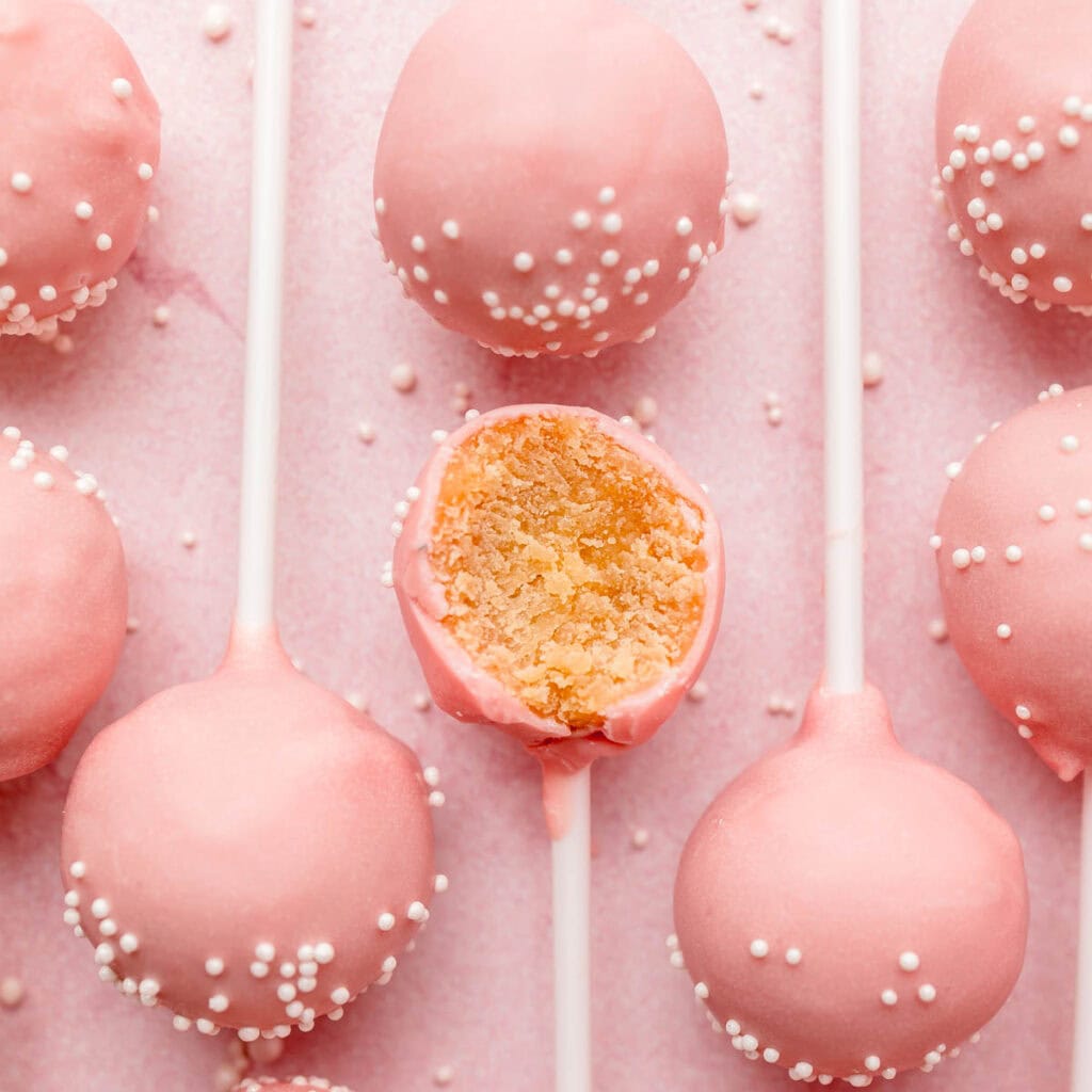 Several gluten-free pink cake pops lined up on pink surface, one with bite taken.