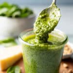 Basil pesto sauce dripping from spoon back into jar filled with pesto sauce.