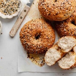 A close-up shot of a gluten-free bagel with a crispy, golden crust covered in everything bagel seasoning, with a knife and small bowl of seasoning nearby. One bagel is cut open to reveal the airy interior.