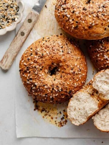 A close-up shot of a gluten-free bagel with a crispy, golden crust covered in everything bagel seasoning, with a knife and small bowl of seasoning nearby. One bagel is cut open to reveal the airy interior.