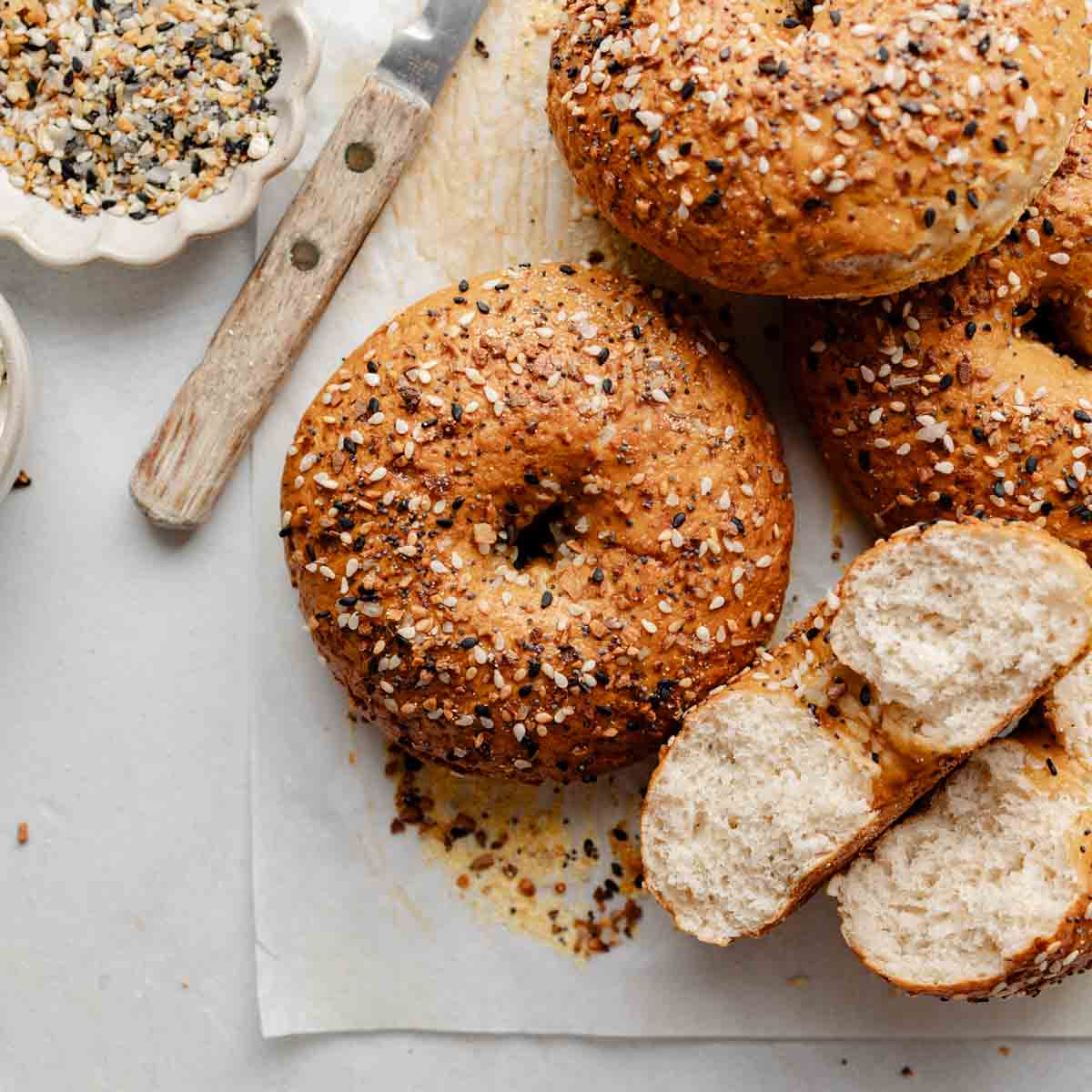 A close-up shot of a gluten-free bagel with a crispy, golden crust covered in everything bagel seasoning, with a knife and small bowl of seasoning nearby. One bagel is cut open to reveal the airy interior.